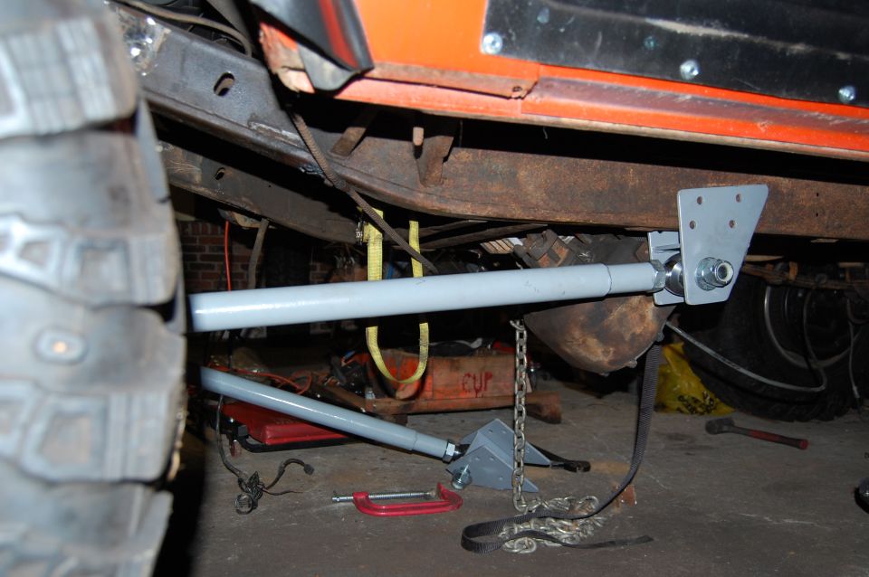 Undercarriage of an orange vehicle with large tire, rusty frame, and suspension components. A "SUPERMOTORS.NET" watermark is visible