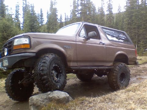 1993 Ford Bronco - brown lifted Ford Bronco with oversized mud tires parked on a rocky forest trail among pine trees.