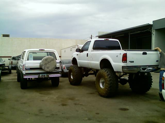 White Ford Bronco with spare tire on back, parked next to a lifted white Ford F-Series truck