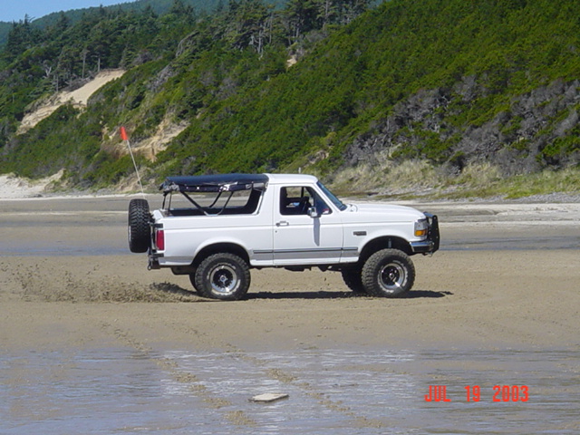 White Ford Bronco with a spare tire on the back driving on a beach with dunes and trees in the background