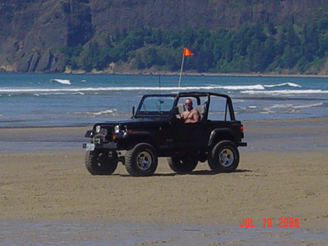Black Jeep Wrangler with a man driving, parked on a sandy beach near the ocean. Orange flag on a pole attached to the Jeep