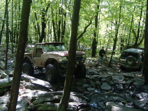 1988 Ford Bronco - Two off-road vehicles navigate a challenging rocky path through a dense forest.