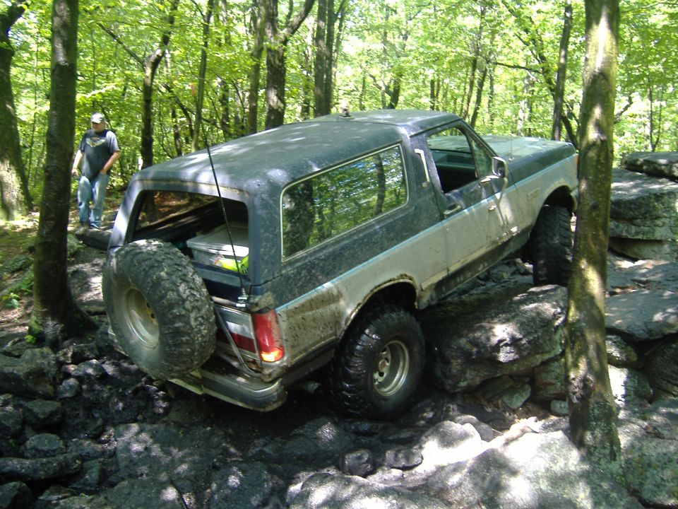 Ford Bronco climbing over rocks on a wooded trail. Spare tire mounted on the back