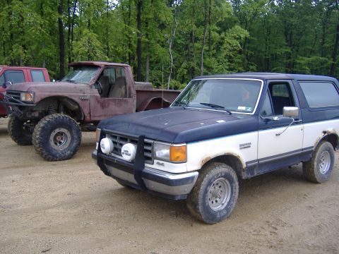 1988 Ford Bronco - A two-tone Ford Bronco is parked on a dirt road with a lifted pickup truck beside it.