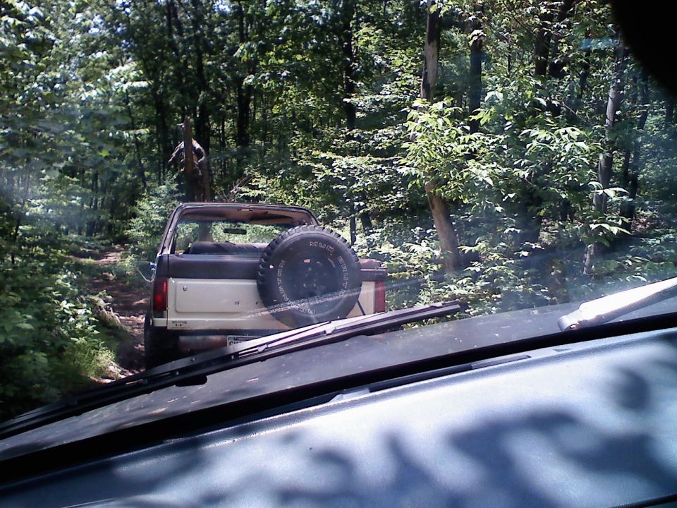 Brown and tan Ford Bronco with spare tire on rear, driving on dirt path in woods