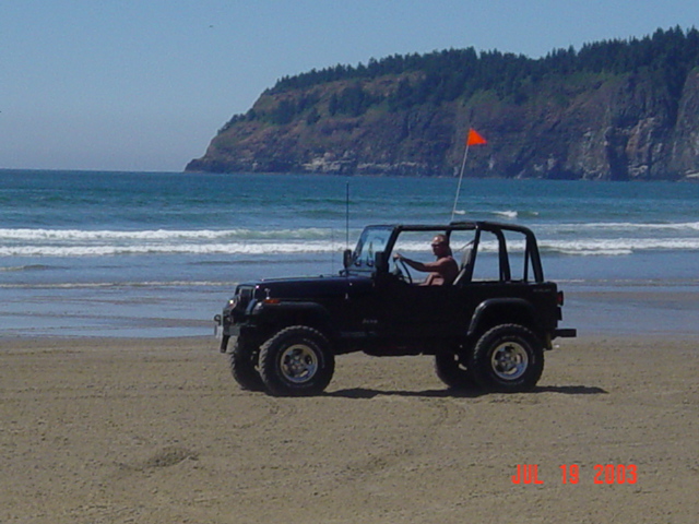 Topless dark Jeep Wrangler YJ driving the surf line below a forested headland