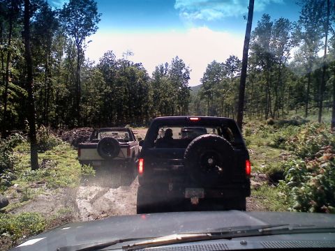 1988 Ford Bronco - Two SUVs navigate a wooded off-road path, kicking up dust.