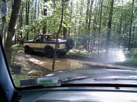 1988 Ford Bronco - A two-tone brown and white Ford Bronco navigates a muddy trail in a wooded area.