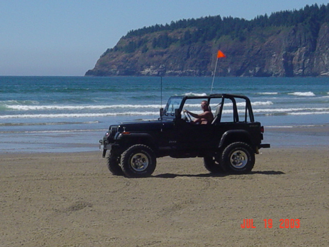 Black 1995 Jeep Wrangler driving on a beach with ocean and a hill in the background
