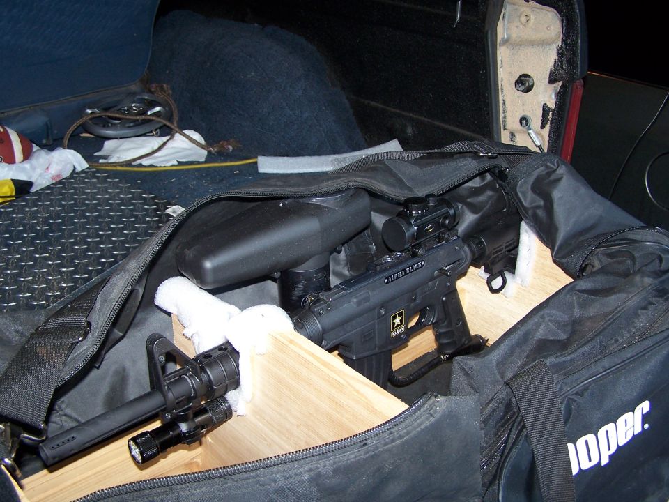 Black tactical paintball gun with scope and flashlight in a duffel bag inside a vehicle