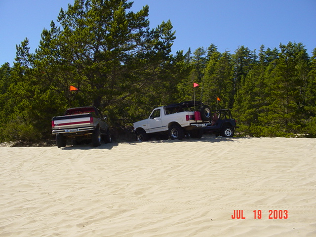 Three off-road vehicles on sand dunes, one is a white pickup truck, another a dark SUV, and a third a dark pickup truck