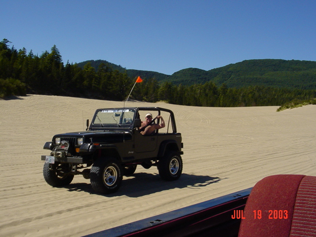 Black Jeep Wrangler driving on sand dunes, front-left quarter view. A person is in the driver's seat. A red seat is visible in the foreground