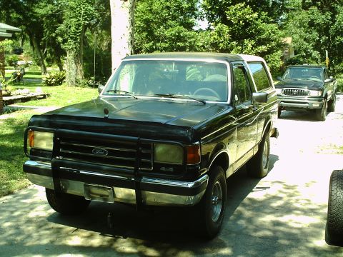1990 Ford Bronco - A black Ford Bronco is parked in a driveway, with a pickup truck visible in the background.