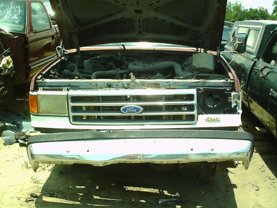 Front view of a 1987 Ford F-150 or Bronco with hood open, showing engine bay. Another truck is parked to the right