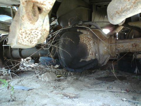 1990 Ford Bronco - The rear axle and suspension of a vehicle are shown, covered in dirt and rust, with dry brush and debris on the ground.