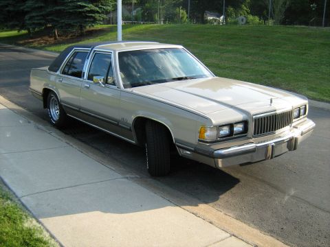 1988 Mercury Grand Marquis - A tan 1988 Mercury Grand Marquis is parked on the side of a road with a grassy hill in the background.