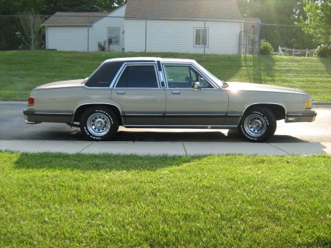 1988 Mercury Grand Marquis - A 1988 Mercury Grand Marquis sedan is seen from the side, parked on a curb with a house and lawn in the background.