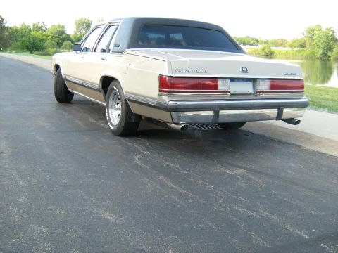 1988 Mercury Grand Marquis - Rear view of a 1988 Mercury Grand Marquis sedan, a cream exterior and a black vinyl roof, parked on an asphalt surface next to a body of wa…