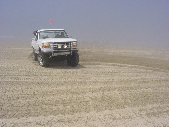 White lifted Ford Bronco with brush guard throwing sand on foggy dunes