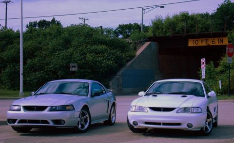 1998 Ford Taurus - Two Ford Mustangs are parked near a bridge with a height restriction sign.