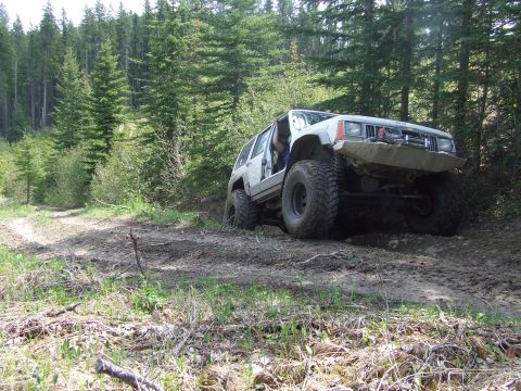 1990 Ford Bronco - Modified Jeep Cherokee XJ navigates a muddy off-road trail through a dense forest.