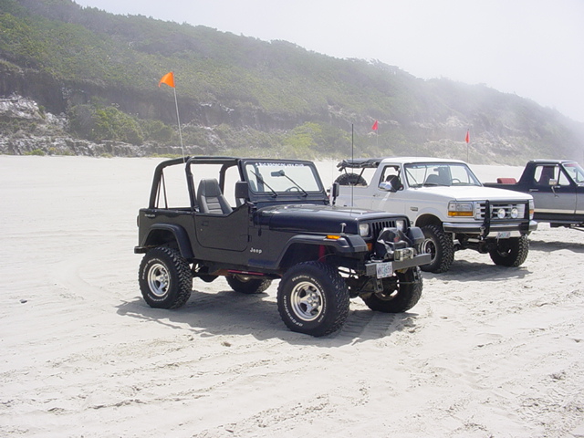 Black Jeep Wrangler and white Ford pickup truck on a sandy beach with hills in the background
