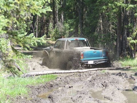 1990 Ford Bronco - A teal pickup truck navigates a challenging muddy off-road course surrounded by dense trees.