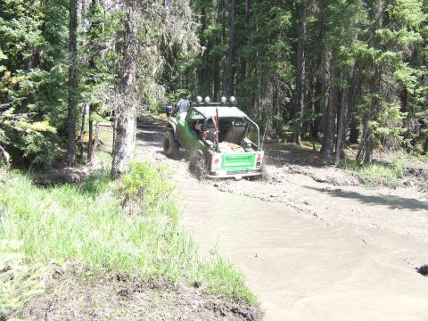 1990 Ford Bronco - A green off-road vehicle navigates a muddy trail surrounded by trees.