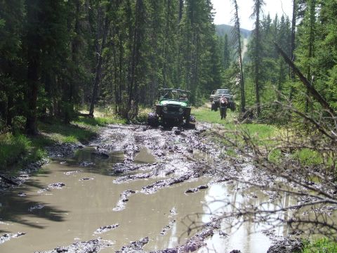 1990 Ford Bronco - Two off-road vehicles navigate a muddy forest trail, with one vehicle deeply stuck in the mire.
