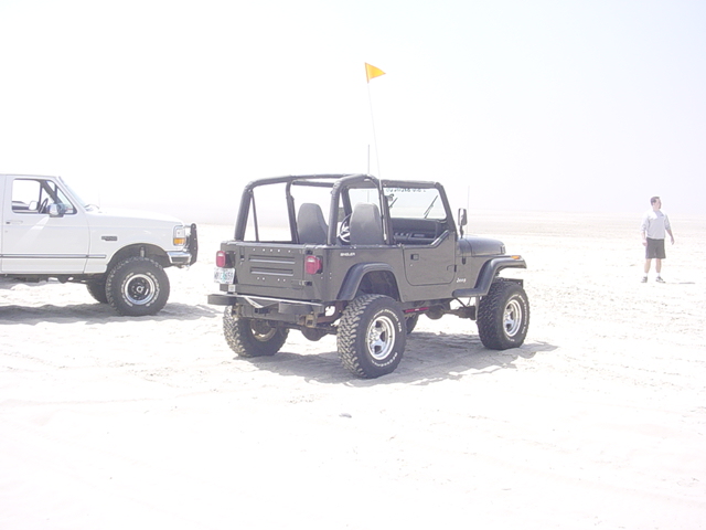 Dark green Jeep Wrangler, rear view, on a sandy beach with a white truck to its left
