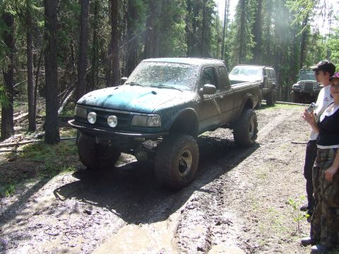 1990 Ford Bronco - This modified pickup truck is navigating a muddy trail in a wooded area, with other vehicles and people visible.