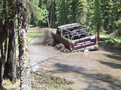 1990 Ford Bronco - An off-road vehicle navigates a muddy trail, kicking up water and mud as it goes.