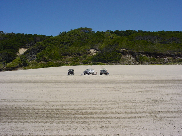 Three SUVs parked on a wide sandy beach with green hills in the background