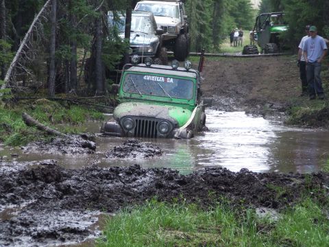 1990 Ford Bronco - Several off-road vehicles navigate a muddy trail, with one Jeep deeply submerged in water.