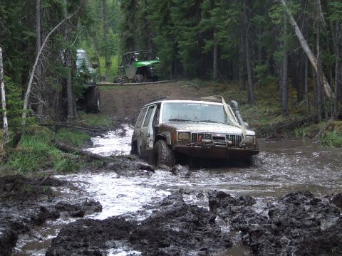 1990 Ford Bronco - Several off-road vehicles navigate a muddy forest trail, with one Jeep deeply mired in water.
