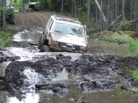 1990 Ford Bronco - A white Jeep Cherokee navigates a muddy trail, with another off-road vehicle visible in the background.