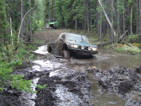 1990 Ford Bronco - Off-road vehicles navigate a muddy trail through a forest.