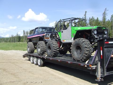 1990 Ford Bronco - Two heavily modified off-road vehicles are transported on a flatbed trailer under a partly cloudy sky.