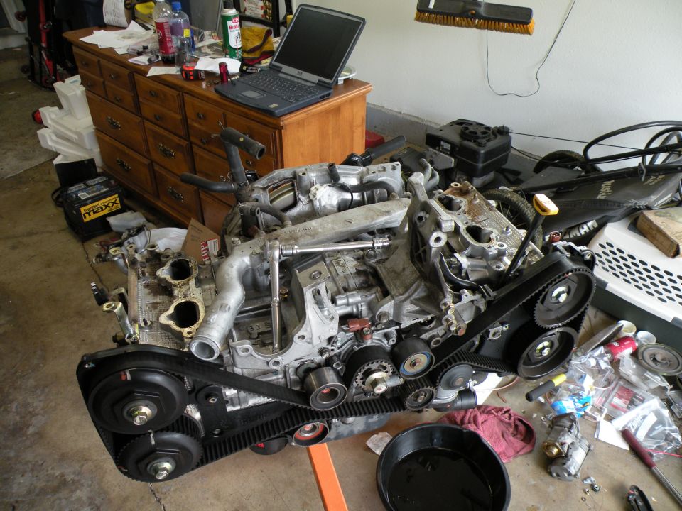Subaru EJ25 engine with timing belt exposed, on an orange engine stand, with tools and parts scattered around