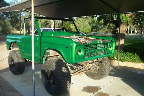 1976 Ford Bronco - A green 1976 Ford Bronco is parked under a canopy, showing its large tires and open hood.