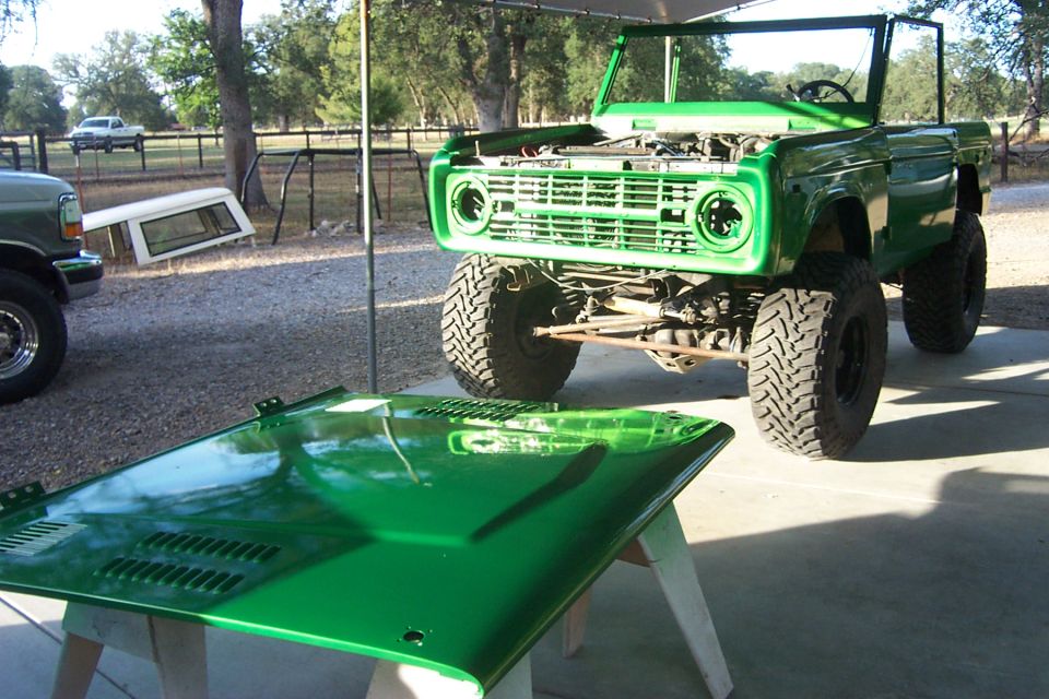 Green Ford Bronco with hood off, sitting outside next to a truck and a camper shell