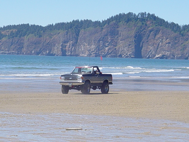 Lifted two-tone Ford Bronco driving on ocean beach with headland behind