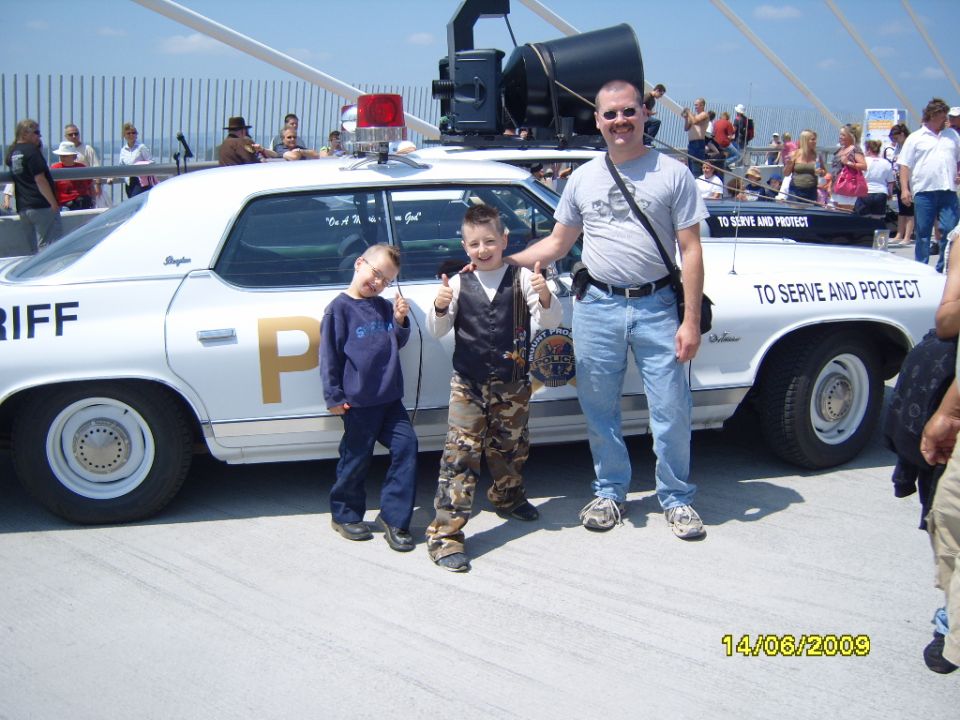 Man and two kids stand next to a white police car with "SHERIFF" and "TO SERVE AND PROTECT" text