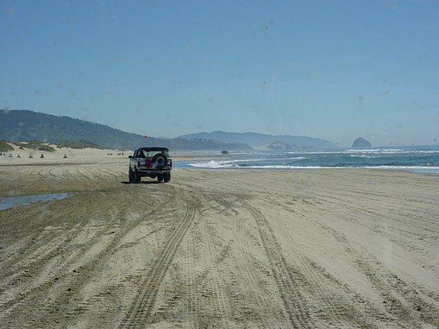1995 Jeep Wrangler driving on a sandy beach with ocean and hills in background