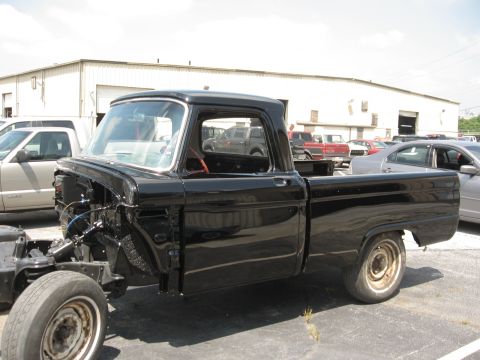 1966 Ford F-100 - A black pickup truck is shown from the side, with its front end disassembled, in a parking lot.