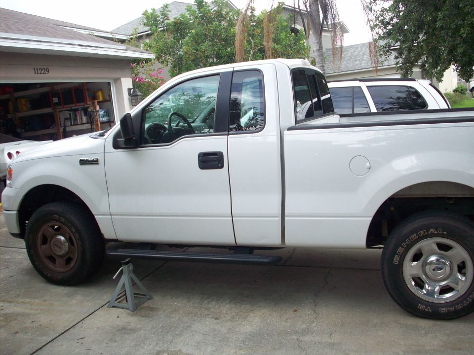 Side view of a white 2006 Ford F-150 pickup truck with one front wheel removed and the truck supported by jack stands. Another white SUV is parked behind it