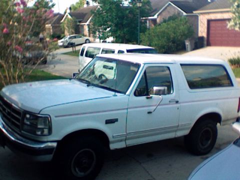 1995 Ford Bronco - A white 1995 Ford Bronco is seen parked in a suburban driveway, with other vehicles visible in the background.