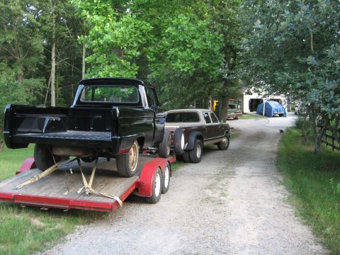 1966 Ford F-100 - A black pickup truck is loaded onto a trailer, with another pickup truck parked behind it on a gravel driveway.