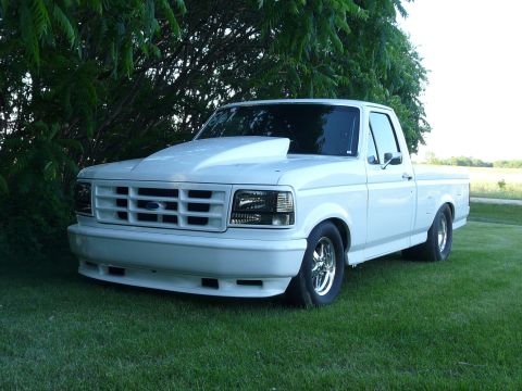 1994 Ford F-150 - Three-quarter front view of a lowered white Ford regular-cab short-bed pickup parked on grass under a tree at dusk, wearing a large cowl ho…