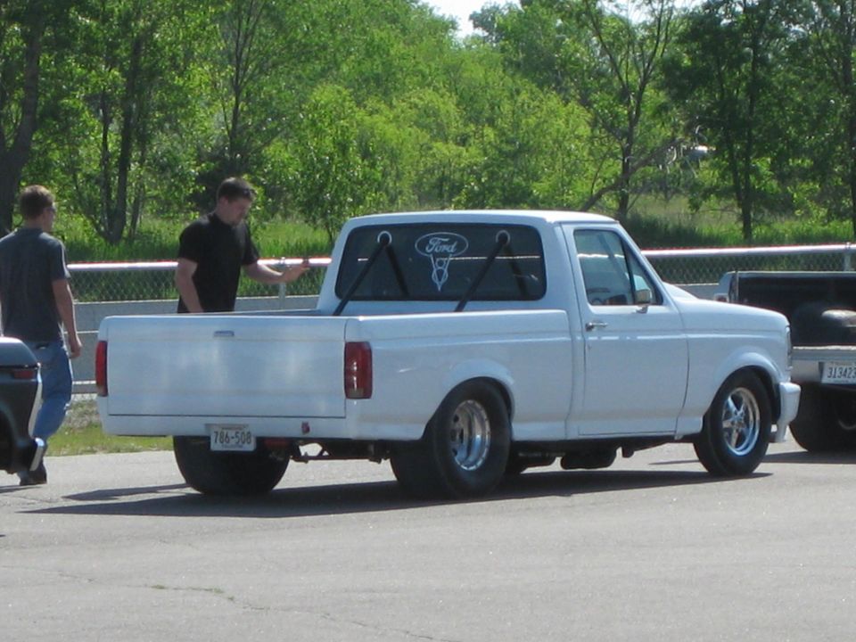 Lowered white Ford pickup with drag rear tires in a track pit area, rear three-quarter view